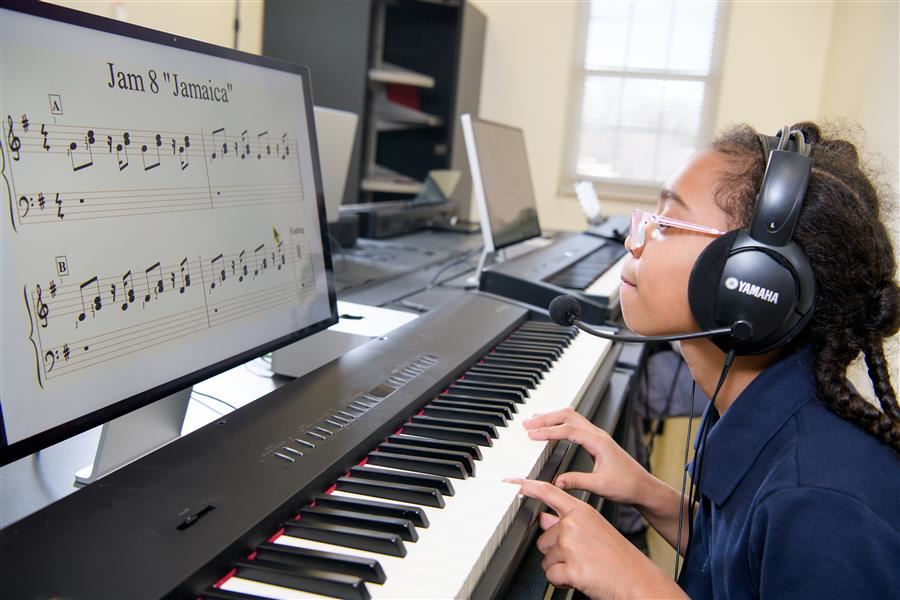 A student looks up at a screen displaying Jam 8 "Jamaica" while playing the piano. She wears a headset.
